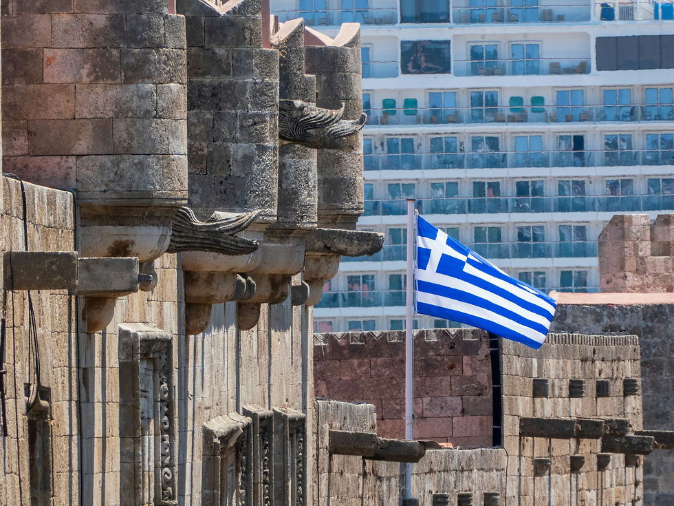 A close-up view of stone walls and dragon gargoyles in the Medieval Old Town of Rhodes, featuring a Greek flag on a flagpole with a large cruise ship anchored in the port behind it.