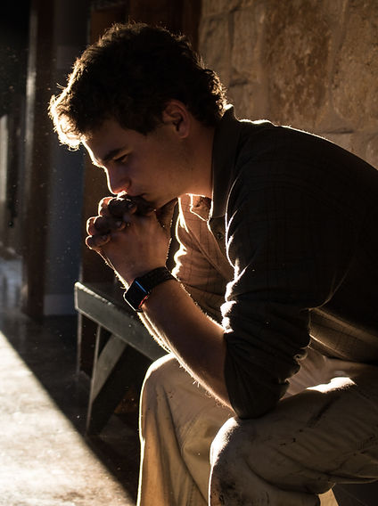 Young man sits in profile, hands clasped near his mouth, in warm backlight by a stone wall, looking pensive and reflective.