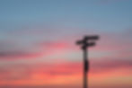 A sunset scene with a blue sky to the top of the image, with streaks of red towards the horizon as the Sun sets.  In the foreground thre is a signpost with the direction arrows pointing in all directions.