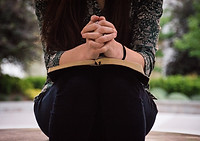 A picture of a woman sitting on a ledge with an open bible in her lap and her hands folded in prayer above the bible.  We do not see the woman's head or face in the picture.