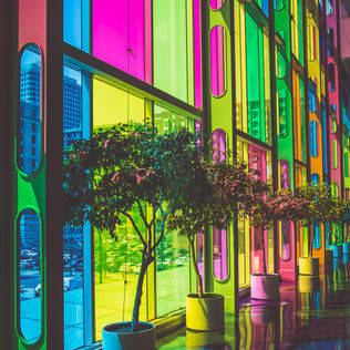 Colorful windows cast rainbow hues on potted trees and a glossy floor in a bright urban setting. Buildings are visible outside.