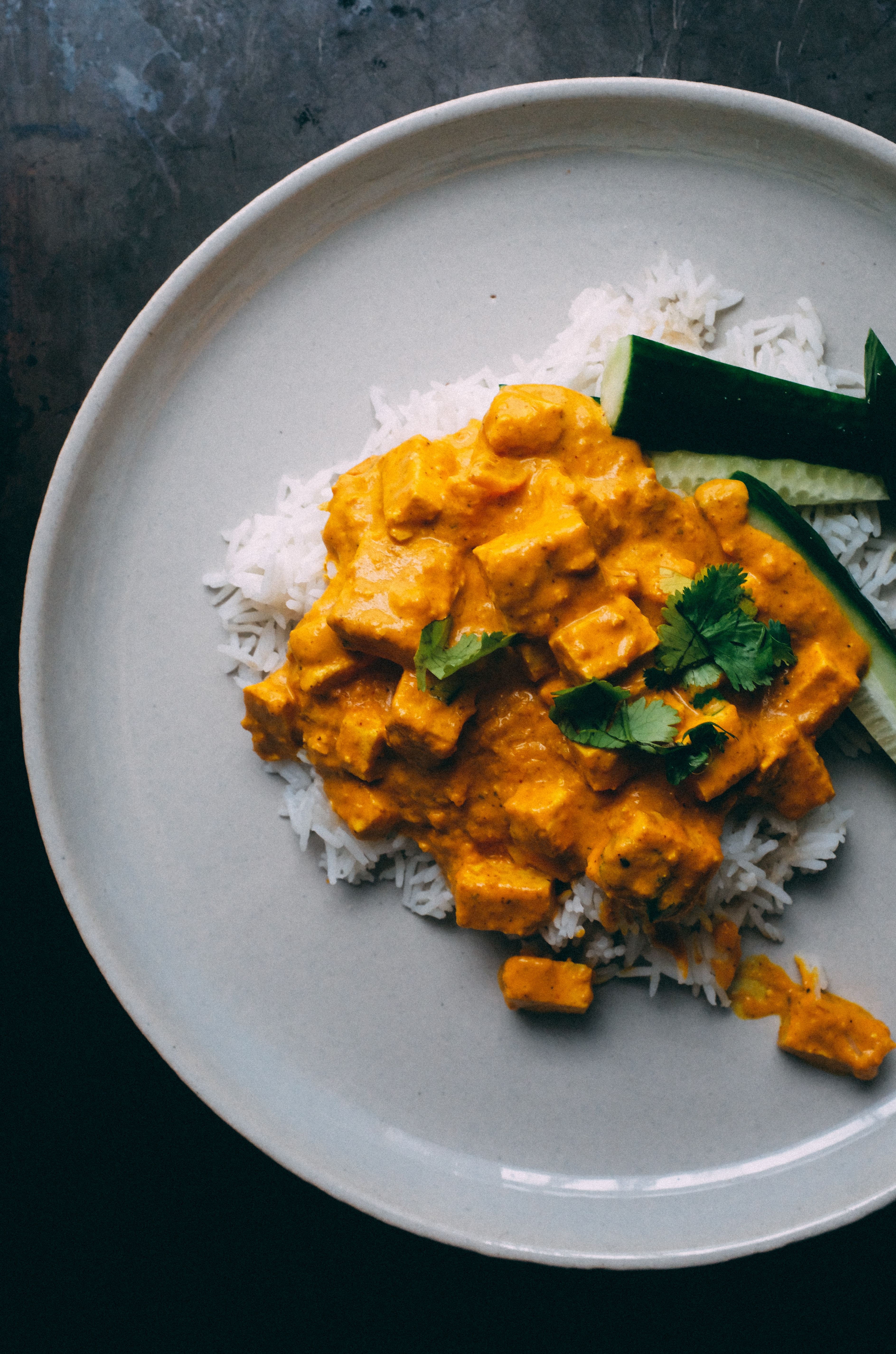 Plate of white rice topped with vibrant yellow tofu curry, garnished with cilantro and cucumber slices on a neutral background.