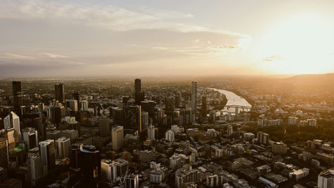 Aerial view of a cityscape at sunset, tall buildings, river winding through. Warm glow and hazy sky create a serene atmosphere.