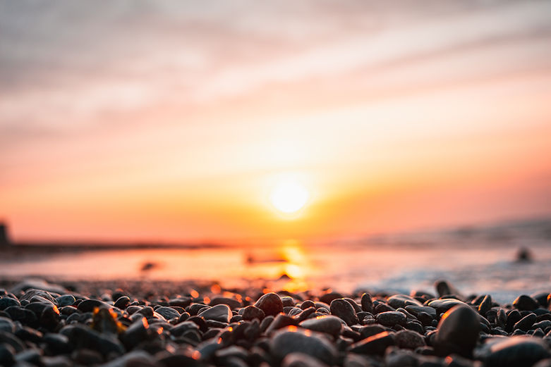 Background Image - pebbles on a beach with the sun reflecting over the sea.