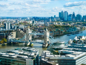 Aerial view of Tower Bridge in London on a sunny day, with cityscape and river in the background. Skyscrapers visible under a blue sky.