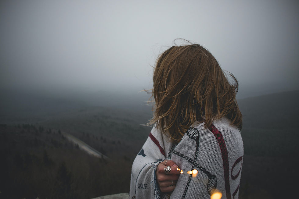 A person wrapped in a blanket stands overlooking a foggy, expansive landscape, holding delicate string lights that softly illuminate the scene.