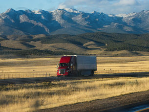 Red truck on a highway through a golden field, with majestic snow-capped mountains in the background under a blue sky. Mood is serene.