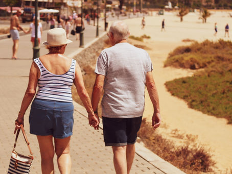 Elderly couple holding hands, walking on a sunny beach promenade. Woman carries a striped bag. Sand and sparse greenery in the background.