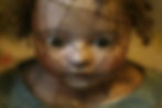 Close-up of a cracked porcelain doll with curly hair and wide eyes, wearing a blue fabric outfit. The mood is eerie and somber.