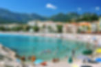 People swim and relax on a sunny beach in the Cote d'Azur with colorful umbrellas, clear blue water, and scenic mountains in the background.