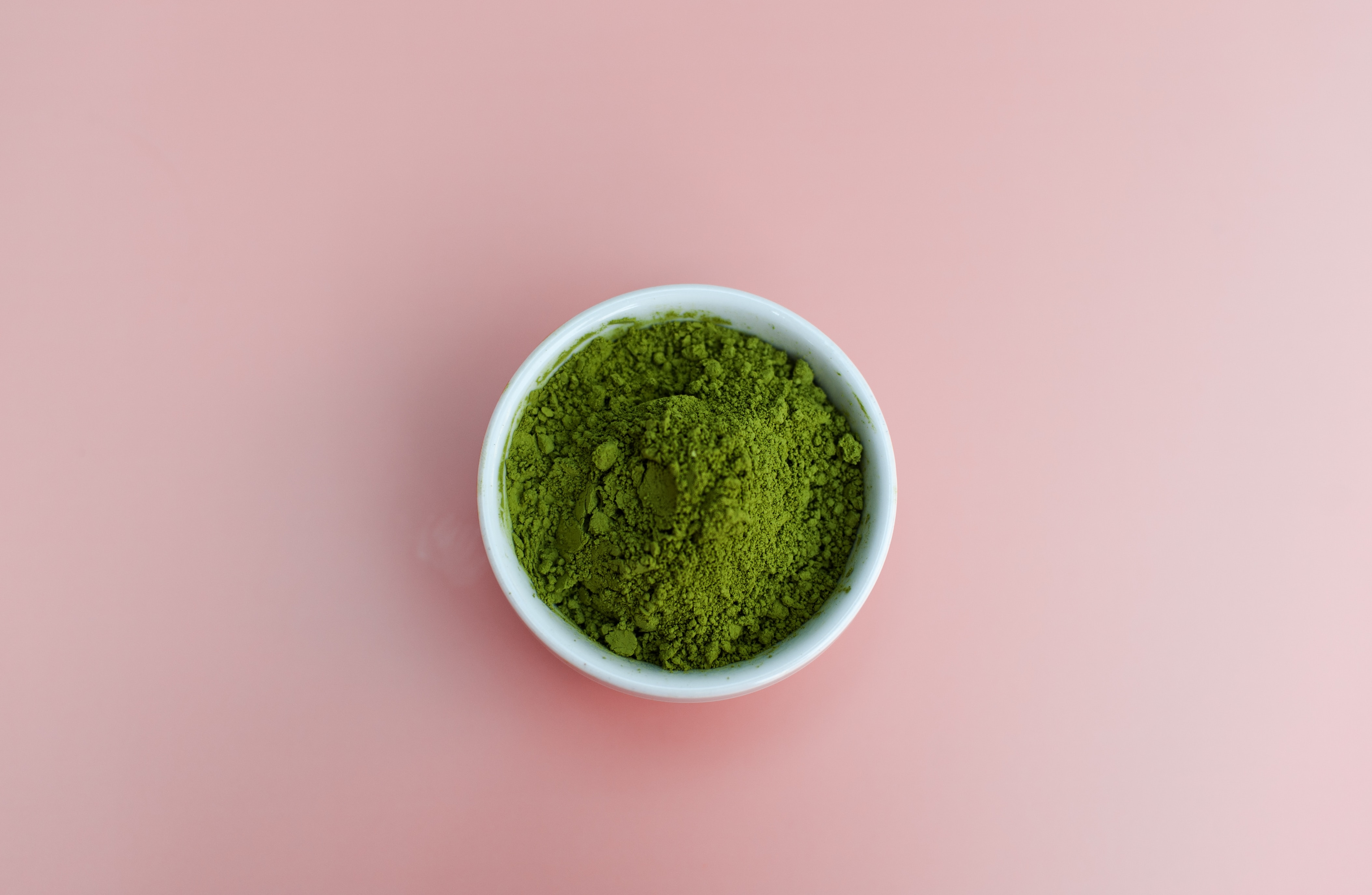 A white bowl filled with green matcha powder sits on a soft pink background, creating a vibrant color contrast.