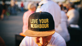 Man wearing a hat with "LOVE YOUR NEIGHBOUR" text sits outdoors among people, creating a friendly, communal atmosphere.