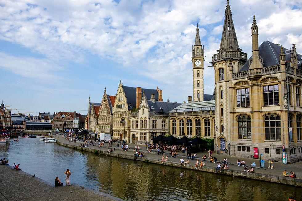 People sit along a canal in Gent with historic buildings, including a tower with a clock. The sky is cloudy, creating a serene atmosphere.