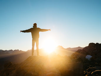 A person stands with arms outstretched as though absorbing the rays of the sun, which is rising in the distance.