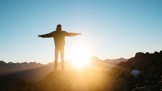 Silhouette of a person with arms outstretched on a rocky mountain at sunrise, clear sky, bright sun behind, creating a serene mood.