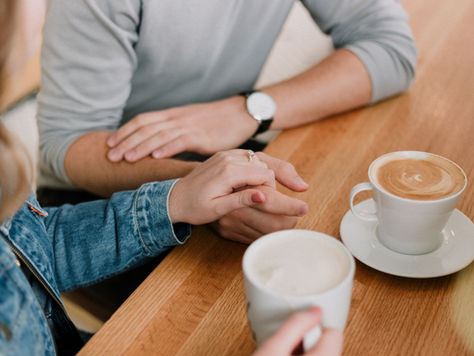 Two people hold coffee mugs over a wooden table. One mug has latte art. The scene conveys warmth and companionship. with blurred lights in the background, creating a warm and intimate atmosphere.