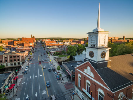 Aerial view of a town with a church steeple on the right, a river, and a bustling street lined with red-brick buildings under a clear blue sky.