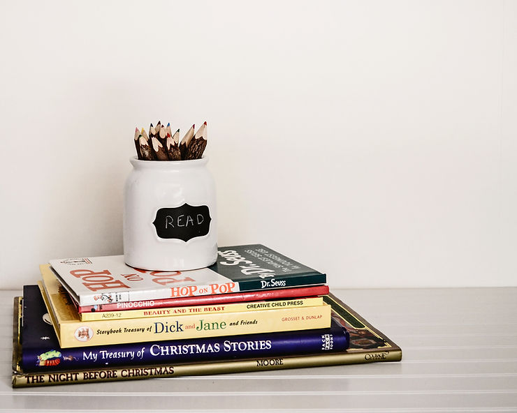 Stack of books on a white shelf, topped with a jar labeled "READ" holding colored pencils. Minimalist setting, neutral tones.
