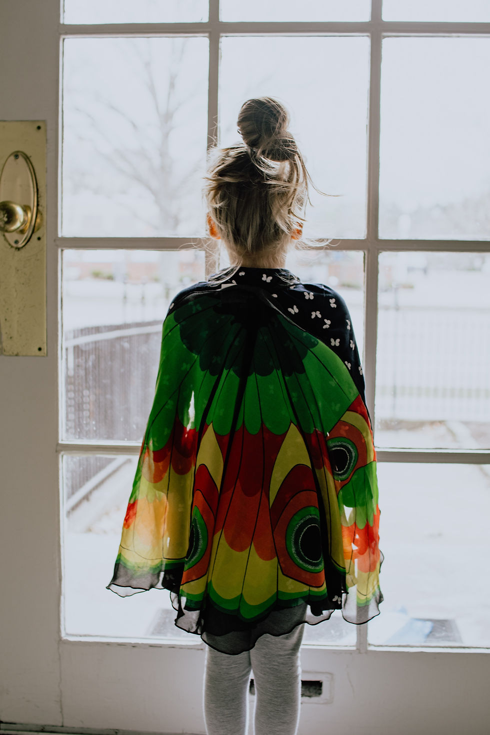 Image of a girl looking out the glass door, wearing a butterfly cape.
