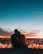 A husband and wife hugging and sitting on a cliff that looks over the city as the sun sets and with candles all around them