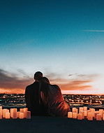 A husband and wife hugging and sitting on a cliff that looks over the city as the sun sets and with candles all around them