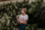 mulher de cabelo loiro e preso com uma trança no topo da cabeça, em frente a várias plantas, vestindo uma blusa que diz: don't look back, you're not going that way (não olhe para trás, você não está indo para aquele lado)