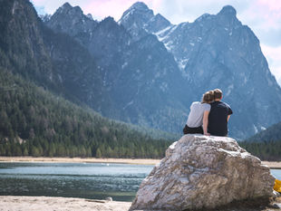 Couple sits on a rock by a lake, facing towering snowy mountains and pine forest under a bright cloudy sky.