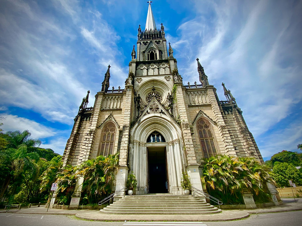 Gothic cathedral with ornate facade and spire, surrounded by palm trees, under a bright blue sky with clouds in Petrópolis. Steps lead to the entrance.