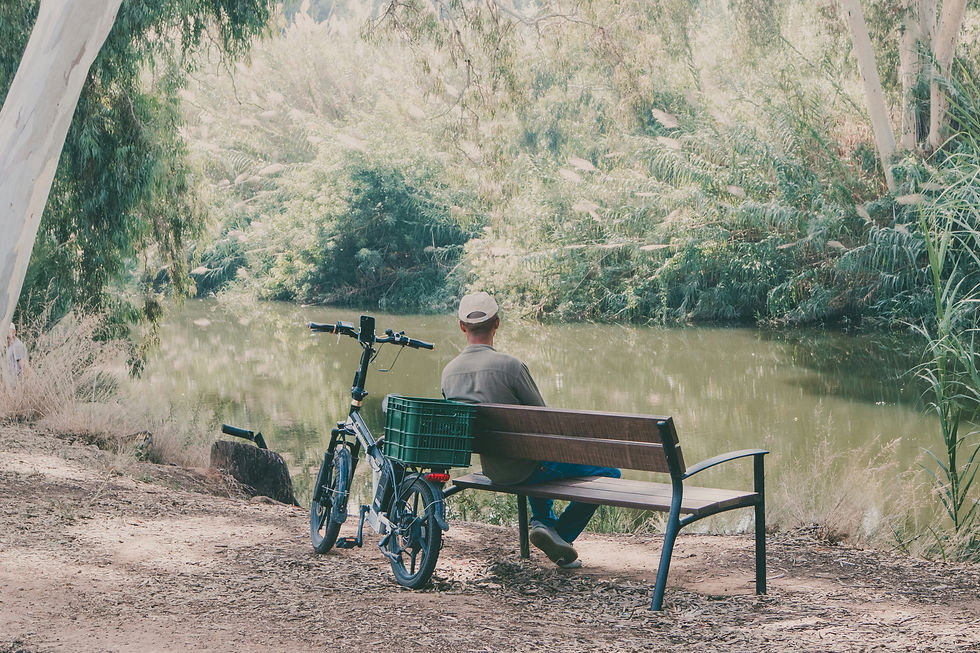 Man sitting on a bench beside a river with a bicycle parked behind him, representing reflection, emotional wellbeing, outdoor activity, and finding calm in nature.