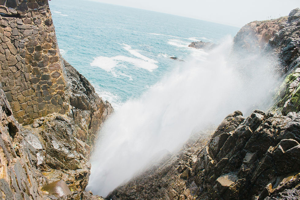 Big ocean wave crashes into rocky seaside cliffs, sending white spray over dark stones and blue water beyond