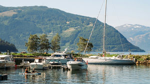 Boats and a sailboat in a calm mountain harbor, with two people paddling on a board under a clear blue sky.