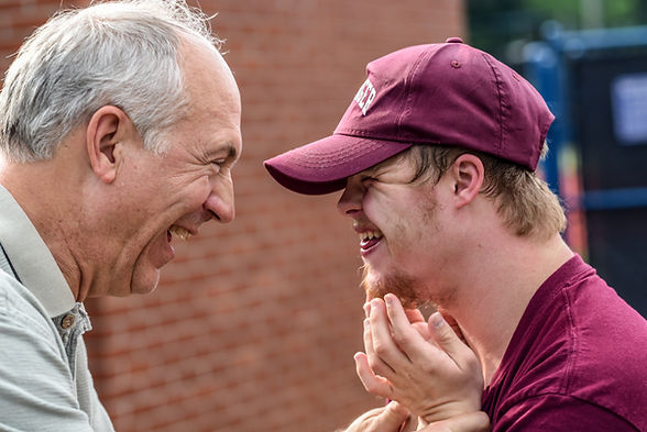 A gentleman with grey hair and a young man with a maroon shirt and hat smilling and laughing together.