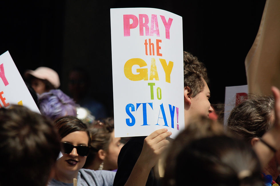 LGBTQ+ pride parade where a multicolored sign says "pray the gay to stay!" held by a white person in a crowd.