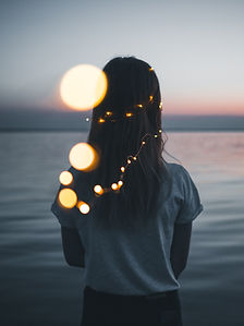 Silhouette of person with fairy lights in hair, facing a calm lake at sunset. Warm bokeh lights create a dreamy ambience.