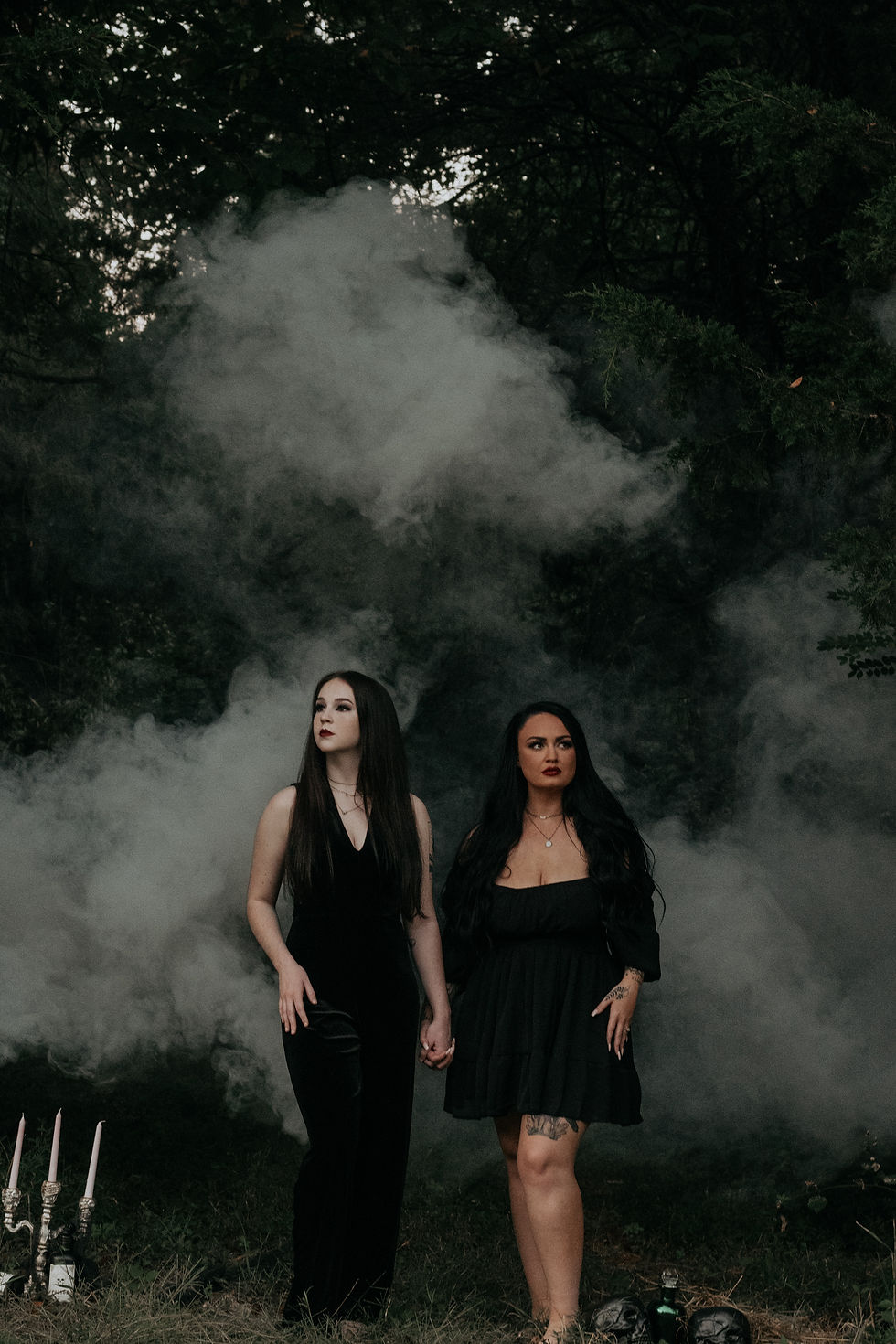Two women in black dresses hold hands in a smoky dark forest, standing amid candles with a moody, gothic atmosphere.