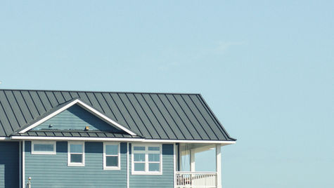 Blue house with white trim and dark roof against clear sky. Balcony visible, American flag in foreground. Peaceful atmosphere.