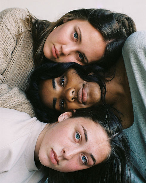 Three teenagers look at the camera, heads together in support-but the image is stacked horizontally so their faces face right and left respectively. They are all girls, one with a beige knitted jumper, she has brown hair and blue eyes and has a mournful expression. Her head is resting on another girl's- a black girl with long black hair, also looking serious but content. Her head is resting on a friend's- she is wearing a white jumper and also has brown hair and blue eyes and has a wistful look