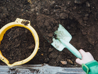 Child’s hand scoops dark soil with a mint shovel beside a yellow bucket in a garden bed, suggesting planting work.