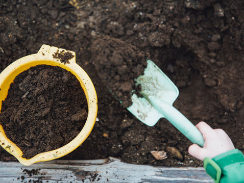 Child’s hand scoops dark soil with a mint shovel beside a yellow bucket in a garden bed, suggesting planting work.