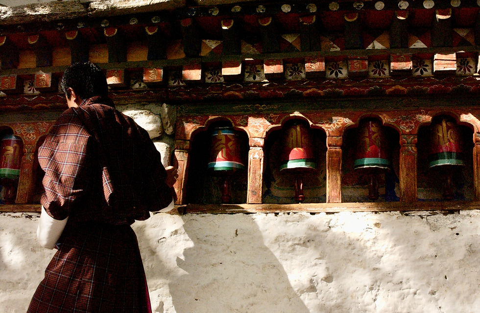 Man in traditional Bhutanese attire spinning prayer wheels at a monastery wall with intricate wood carvings
