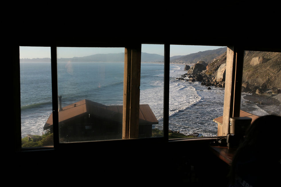 View of a serene sunset over the ocean through a window, framed by curtains. The sky is a mix of blue, orange, and purple hues.