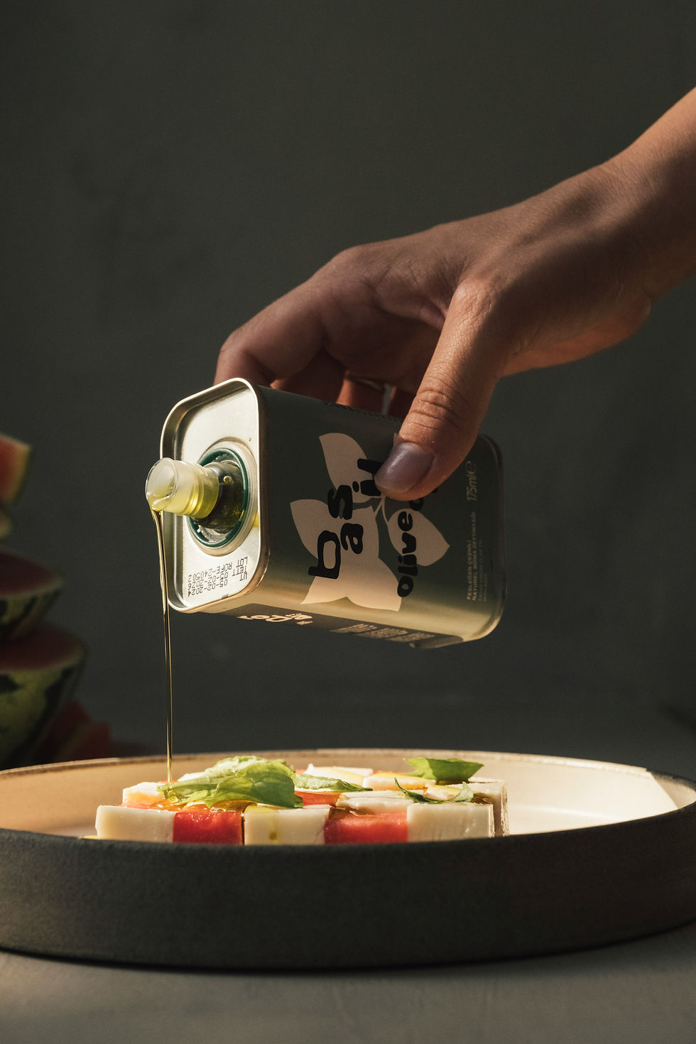 Mano vertiendo aceite de oliva con albahaca sobre ensalada de sandía y queso. Fondo oscuro, luz suave, enfoque en la botella.