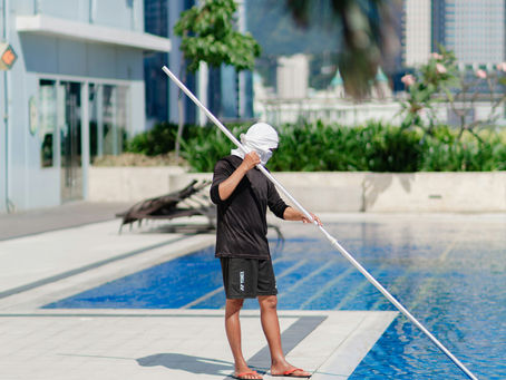 A person in black shorts and shirt cleans a pool with a long pole. Bright sunny day, poolside with greenery and buildings in the background.