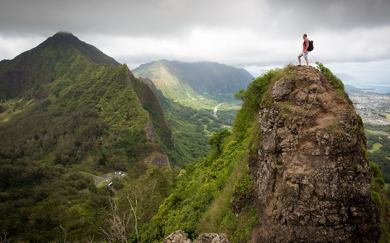 A Hiker on Top of A mountain surrounded by Other Mountains with Green Trees On Them