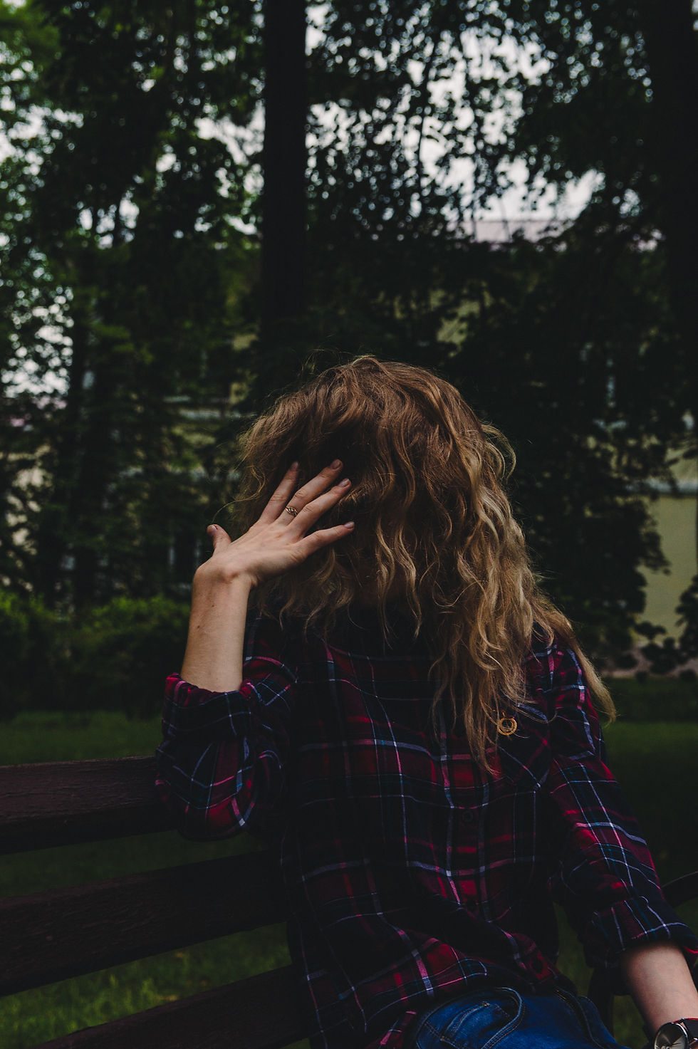 Person with curly hair sitting on a bench in park, wearing a red plaid shirt. Hand raised to head, dark, wooded background.
