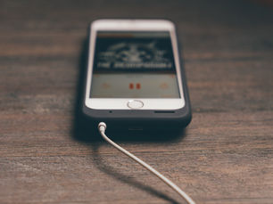 Smartphone on wooden table playing audio, connected with white earphones. Display shows play/pause icon. Minimalist, calm setting.