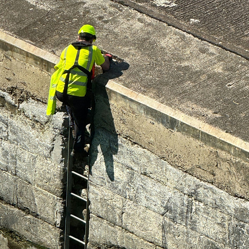 A worker in a neon safety vest and helmet climbs a ladder at the edge of a concrete structure