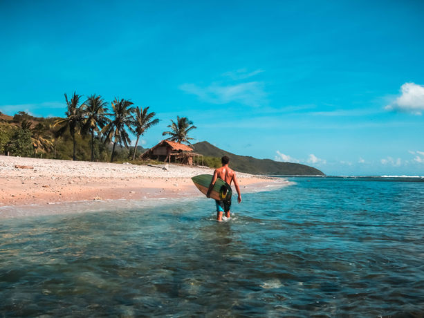 Ambiente balneare e attività ricreative in Indonesia. Una spiaggia quasi deserta, frequentata da un surfista che esce dall’acqua. Sulla terraferma una piccola casa di legno circondata da alcune palme