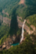 A tall waterfall cascades down a lush, green cliff into a blue pool below. Dense forest surrounds the scene, creating a tranquil atmosphere.