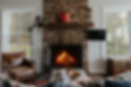 Cozy living room with a lit stone fireplace, leather chair, TV, and vintage trunk beside bright windows.